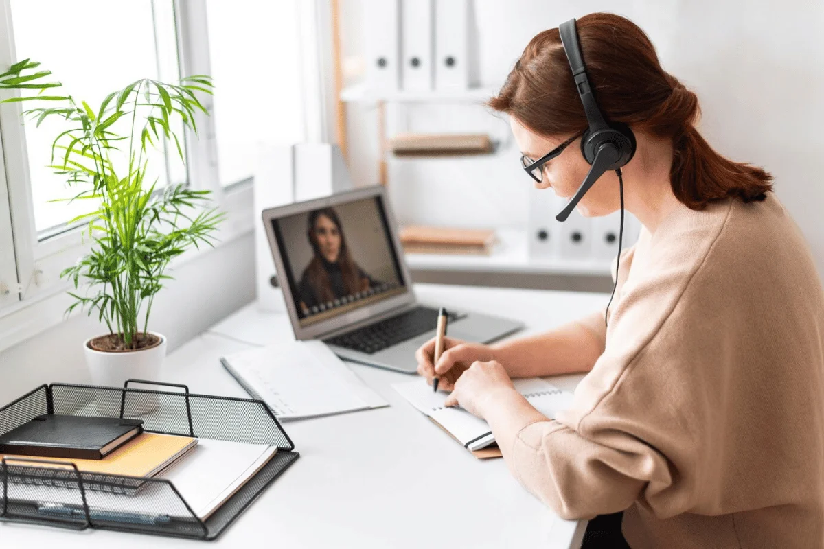 Woman in a virtual meeting taking notes while wearing a headset and using a laptop.