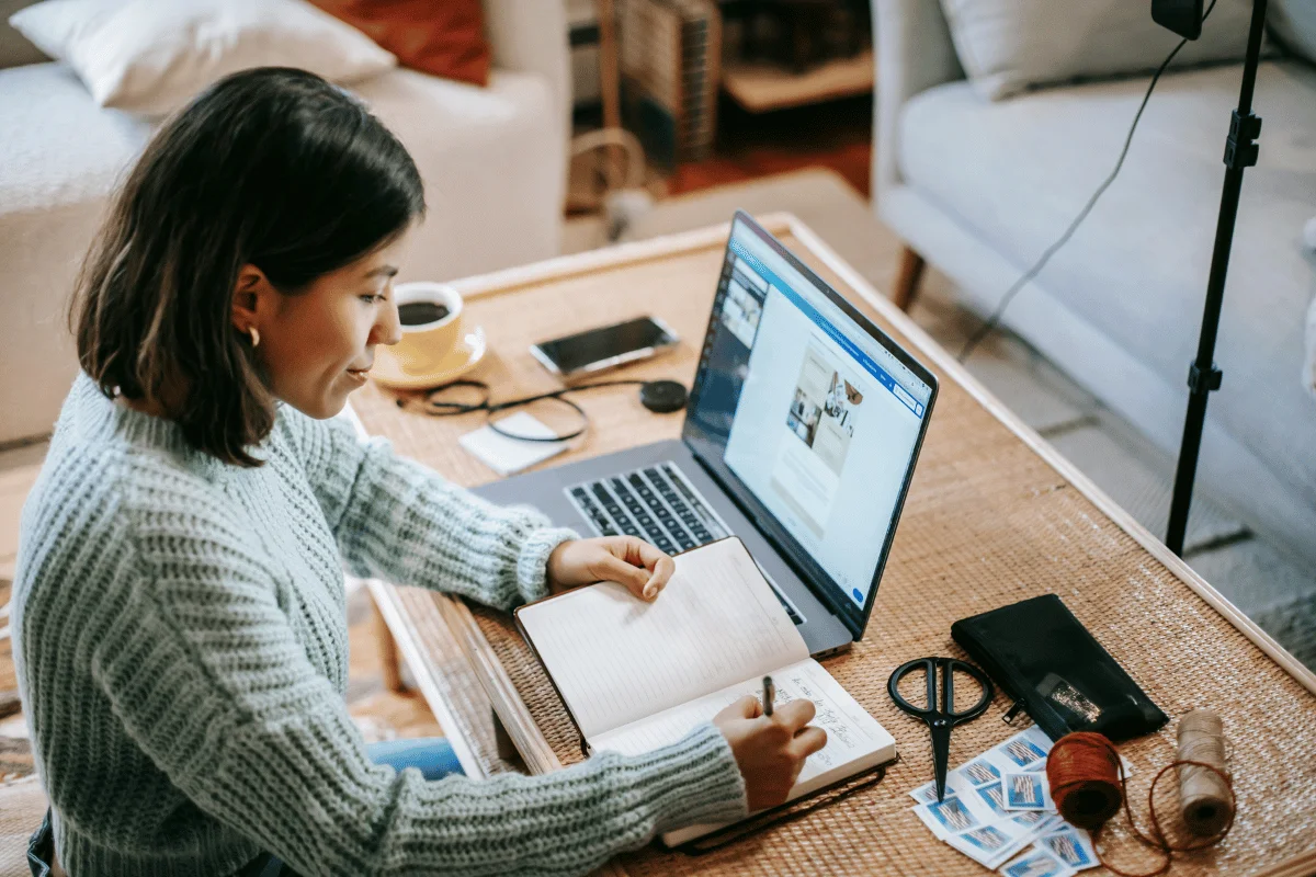 Woman working from home on a laptop with an open notebook, coffee, and scissors on the desk.