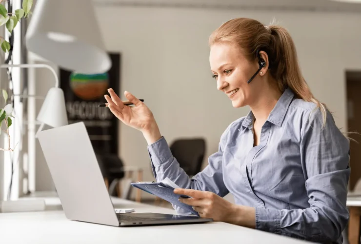 Woman with a ponytail wearing a blue shirt uses a laptop and holds a clipboard, gesturing with a pen during a remote meeting.