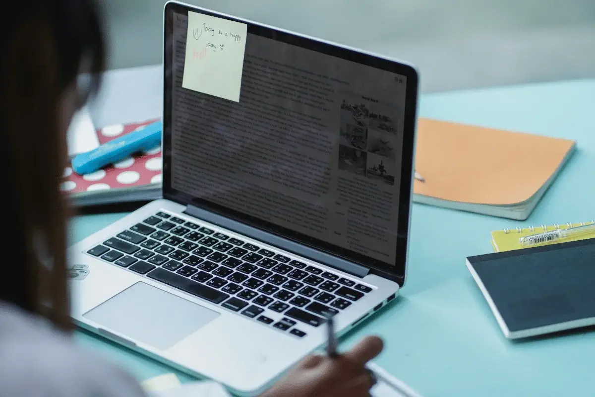 Close-up of a laptop screen with a sticky note and a person writing in a notebook.