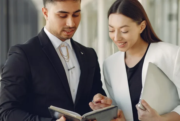 Two professionals reviewing project management documents together in modern office hallway.