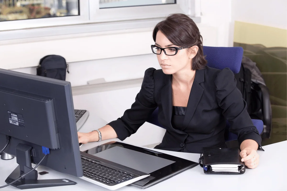 A professional woman works on her laptop and desktop computer at a clean office desk.