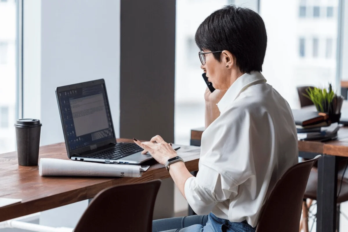 A woman in a white blouse managing work on her laptop while speaking on the phone in an office setting.