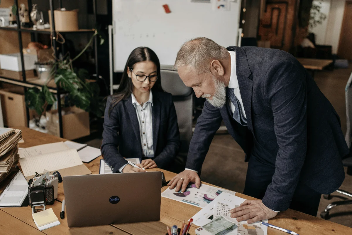 A senior and junior business professional discussing charts and finances at a desk.