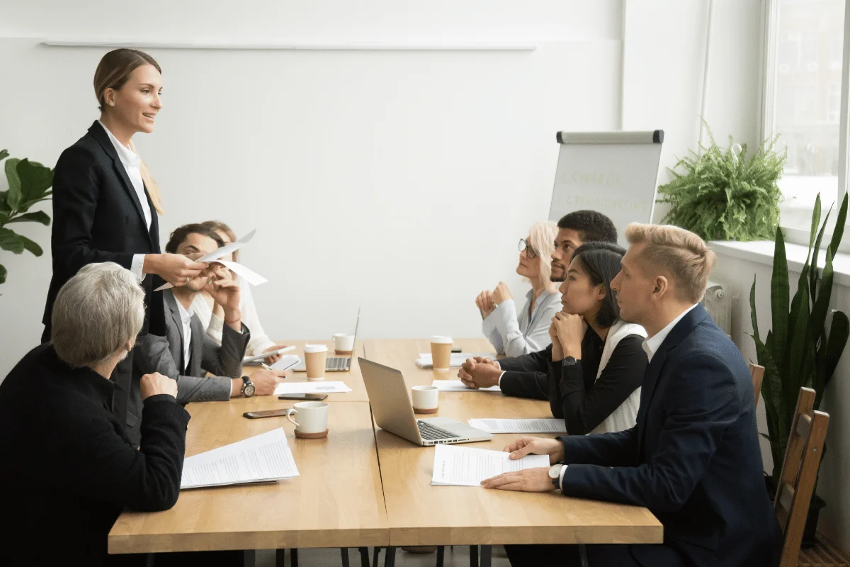A conference room meeting with a group of professionals engaged in discussion and presentations around a wooden table, plants in the background.