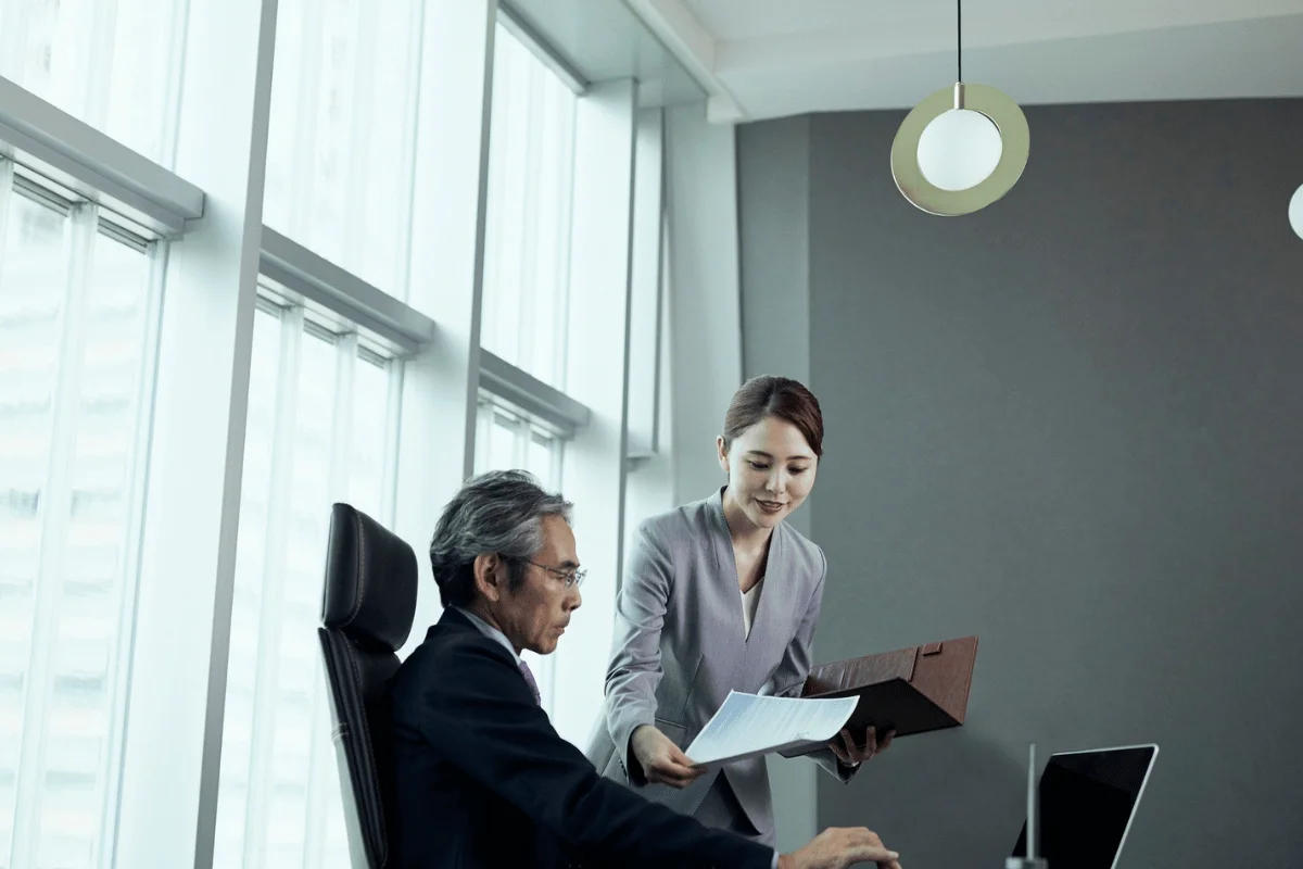Executive working at his desk while a businesswoman hands him a report in a professional office environment.