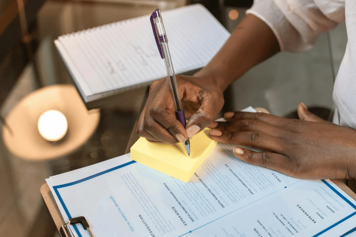 Person writing on sticky notes beside printed resume on clipboard.