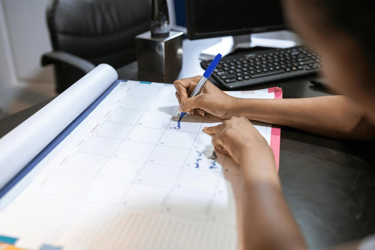Person writing on a monthly calendar with a blue pen, marking down important dates in an office environment.