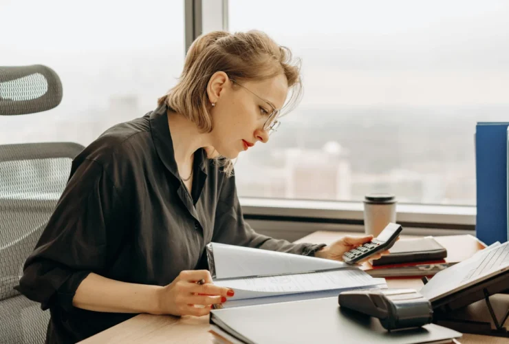 A focused woman in glasses working on financial calculations at a desk with office supplies.