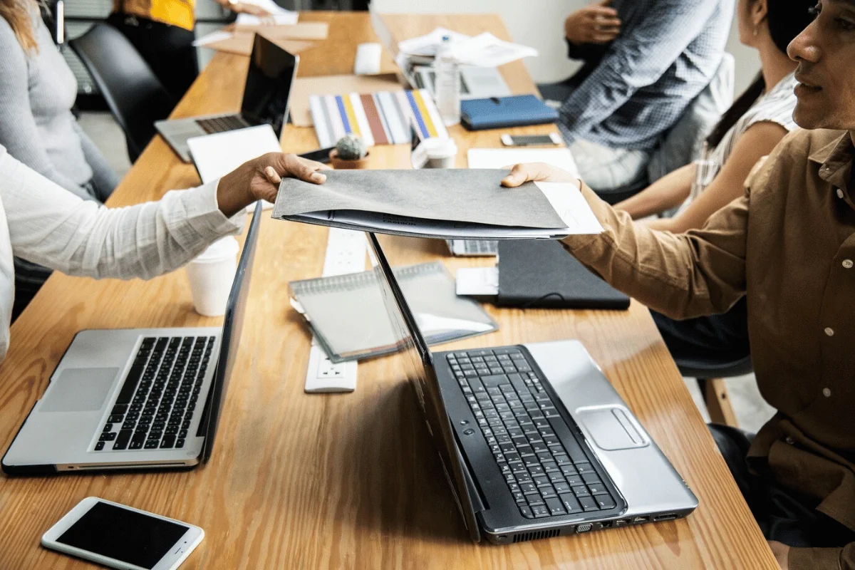 Two professionals exchanging a folder over a desk filled with laptops, notebooks, and office supplies during a business meeting.