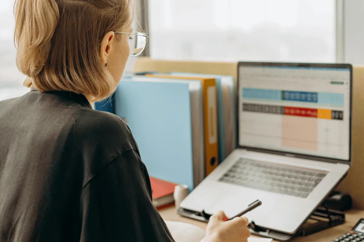 A professional woman analyzing a spreadsheet on her laptop while taking notes in a modern office.