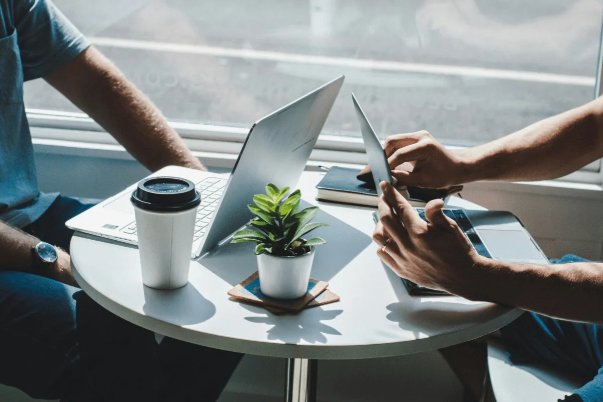 Two professionals working on laptops and a tablet at a small round table near a window, with a coffee cup and a plant in the center.