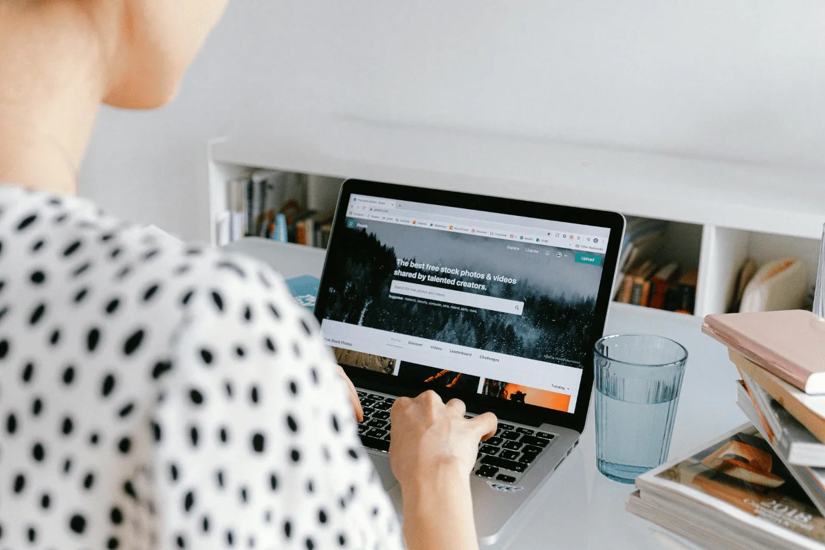 A person types on a laptop displaying a stock photo website, with books and a glass of water on a desk in the background.