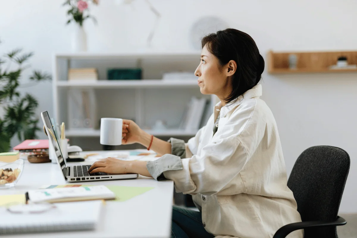 A person sitting at a desk, using a laptop and holding a white mug, surrounded by plants and stationery in a bright workspace.