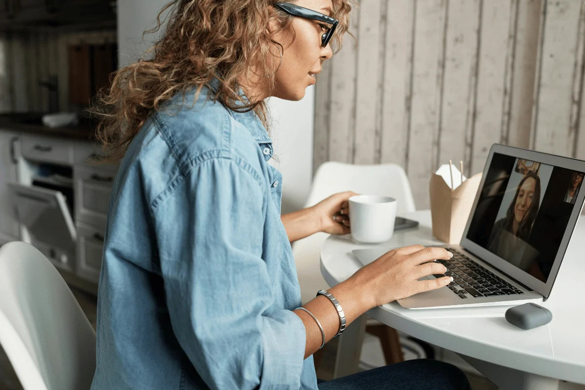 A person sits at a table in a cozy kitchen, holding a coffee cup and using a laptop for a video call.