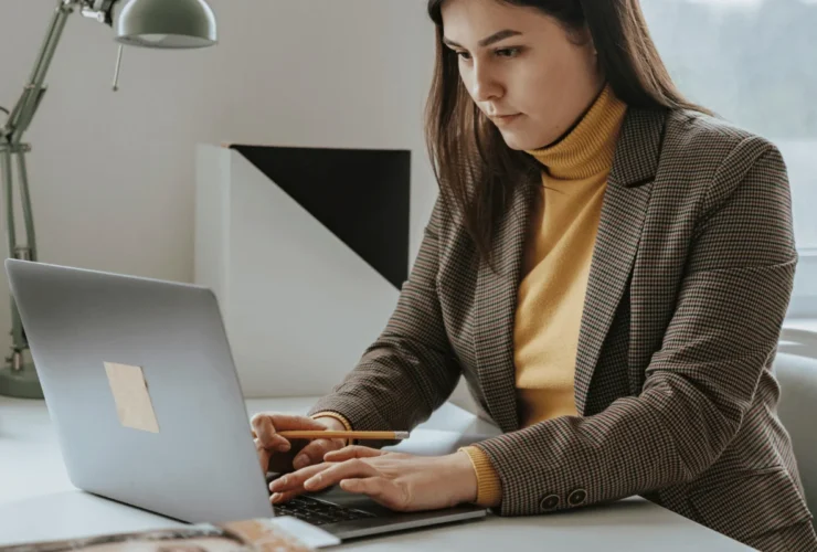 A person in a plaid blazer and yellow turtleneck works on a laptop, taking notes with a pencil at a stylish desk.