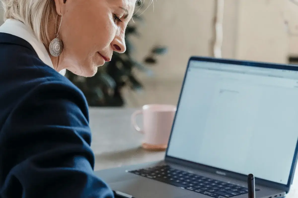 A woman in a blue blazer works intently on a laptop, with a pink mug in the background.
