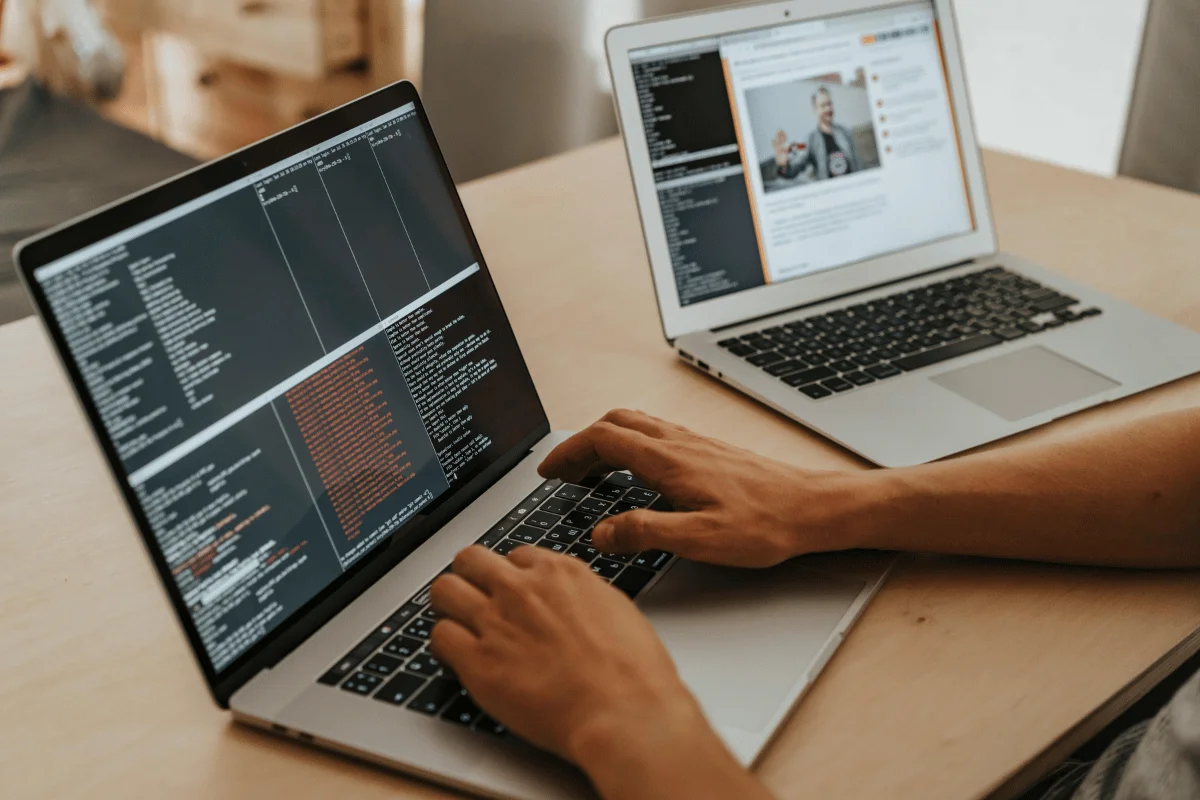 A person coding on a laptop with dark terminal output, while another laptop displays a video conference call and related content.