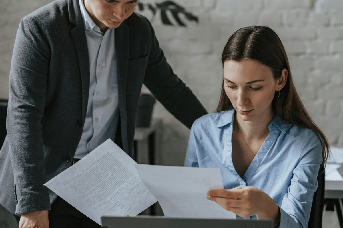 A woman holding papers sits at a desk, while a man in a suit looks over her shoulder, discussing documents in a modern office setting.