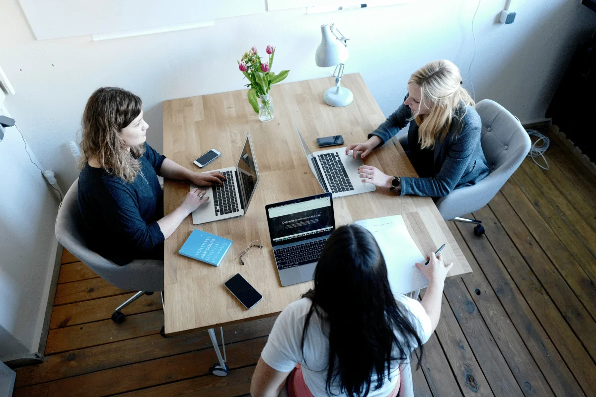 Overhead view of a group of women discussing ideas at a coworking space, with laptops, books, and notes on the table.