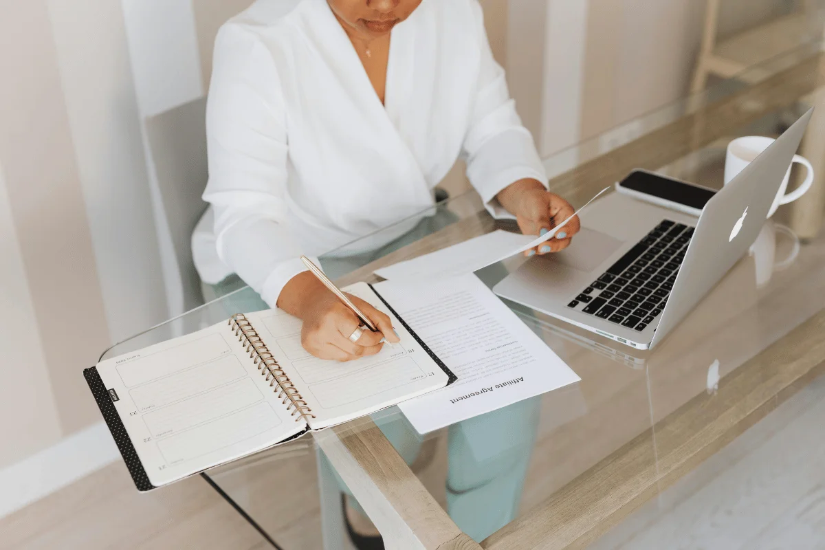 A professional woman in a white blouse reviewing documents and taking notes at a glass office desk.