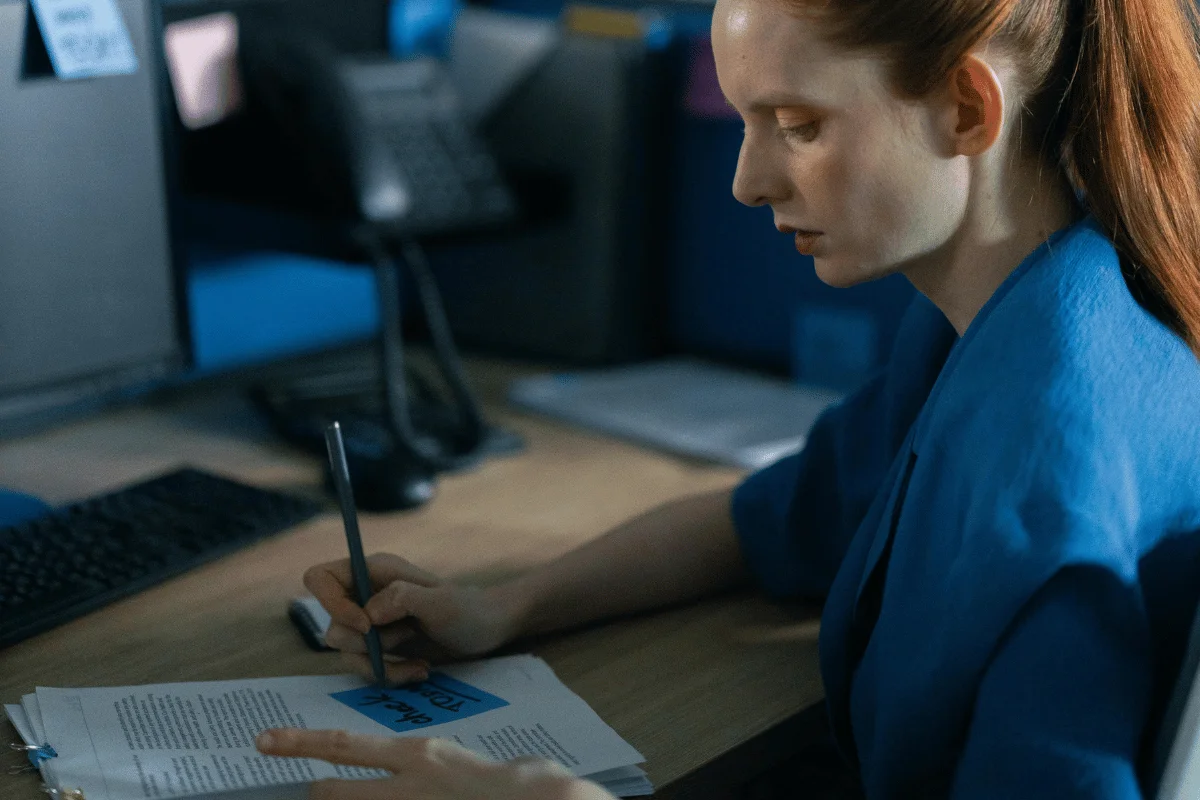 A person in a blue blazer writes notes on a stack of papers at a desk, with a keyboard and office phone in the background.