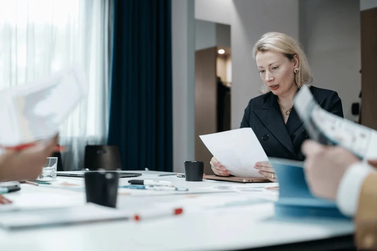 A professional meeting scene featuring a woman reviewing documents at a table, surrounded by colleagues and various stationery items.