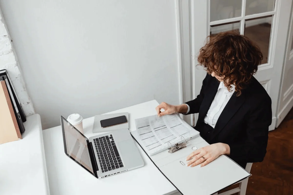 A professional woman in a black suit reviewing documents in a binder at a white office desk with a laptop and coffee.