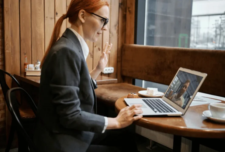 A professional woman in a suit sits at a café table, engaging in a video call on her laptop, with coffee and pastries nearby.