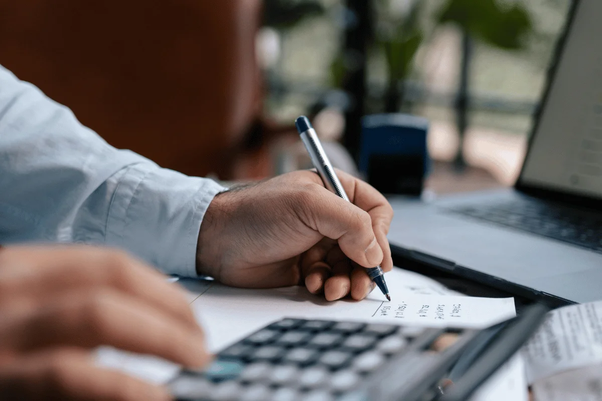 A person in a light blue shirt writes calculations on a paper while using a calculator, with a laptop in the background.