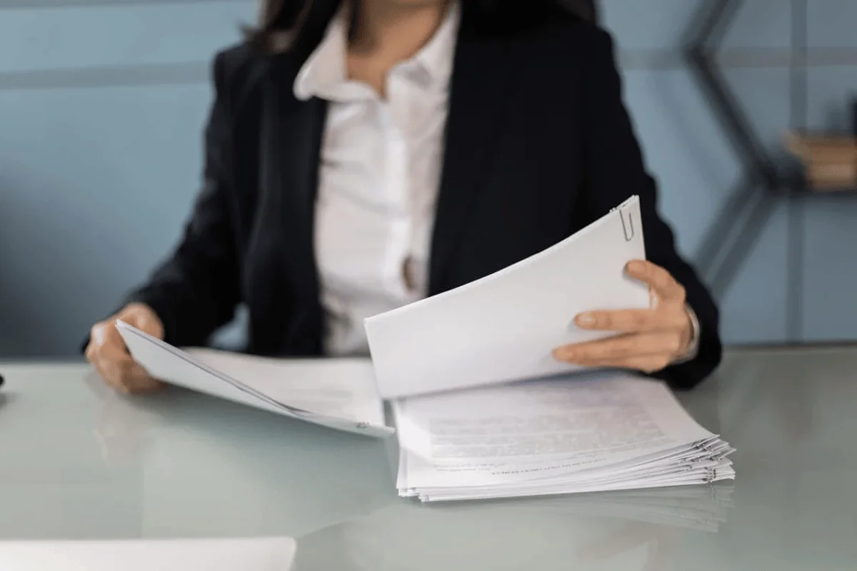 A businesswoman in a black suit reviewing important documents at a glass desk in a modern office.