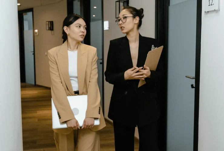 Two professional businesswomen walking and discussing work in a modern office hallway.