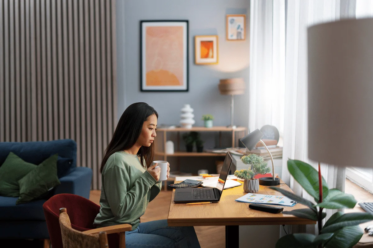 A woman in a cozy workspace sits at a wooden desk, holding a mug, with a laptop and plants around her, surrounded by warm decor.