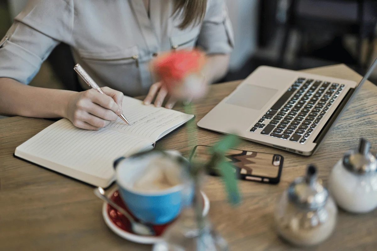 A person writes in a notebook next to a laptop, coffee cup, and flower, capturing a cozy workspace atmosphere.