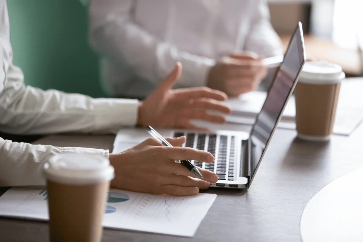 Two professionals engage in a discussion over a laptop and coffee cups, reviewing documents with graphs on a wooden table.