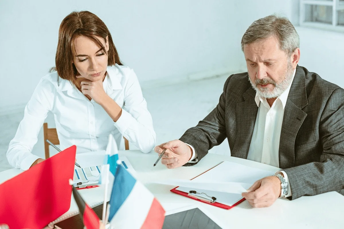 A man and woman in serious discussion at a table with international flags, focusing on documents.