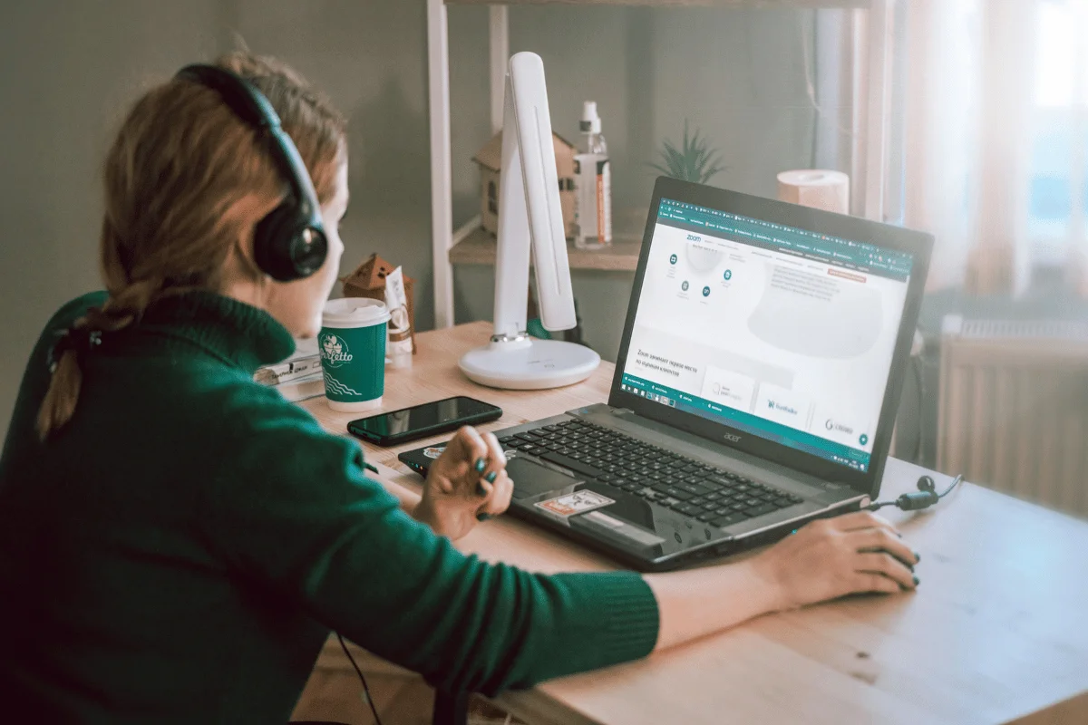 A woman in headphones sits at a desk with a laptop, smartphone, and coffee, focused on a Zoom webpage. 