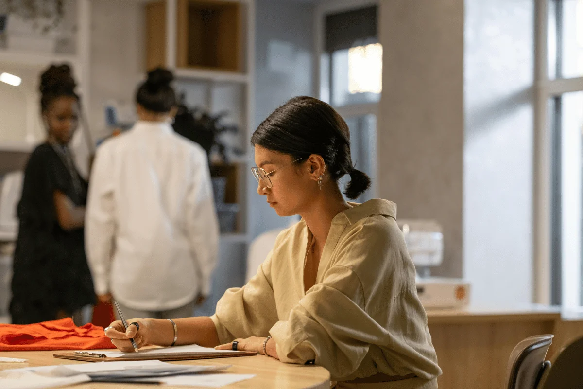 A woman in a beige shirt writes notes at a table, while two individuals converse in the background, surrounded by a modern workspace.