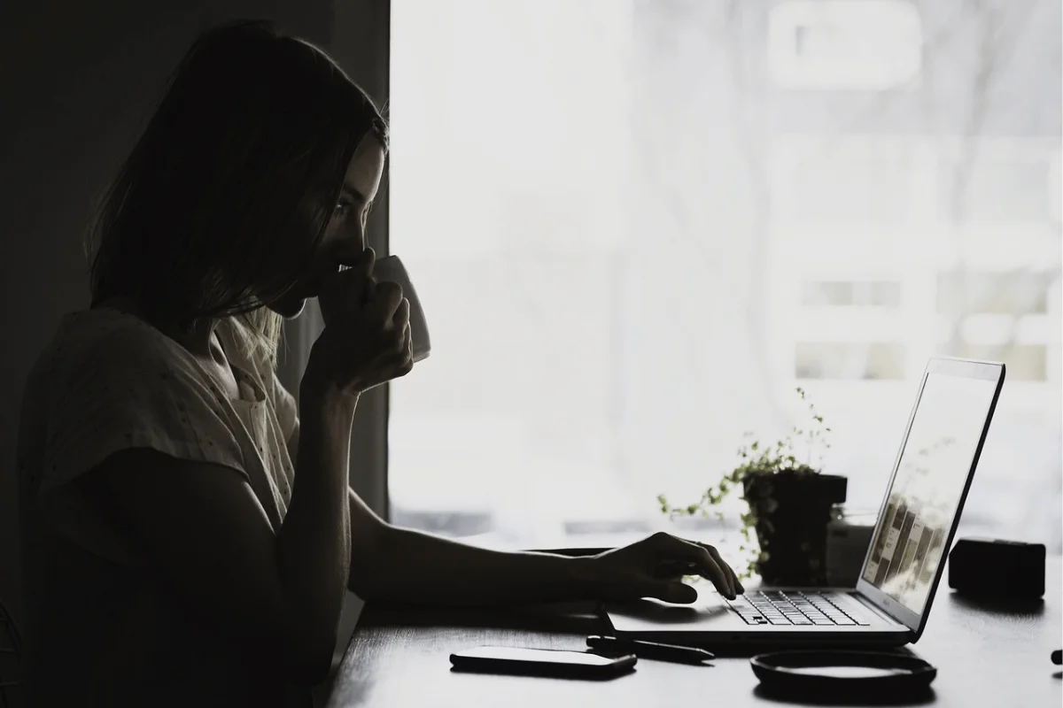 A person sits at a desk with a laptop, thoughtfully sipping from a cup, with a small plant and pens nearby, illuminated by natural light.