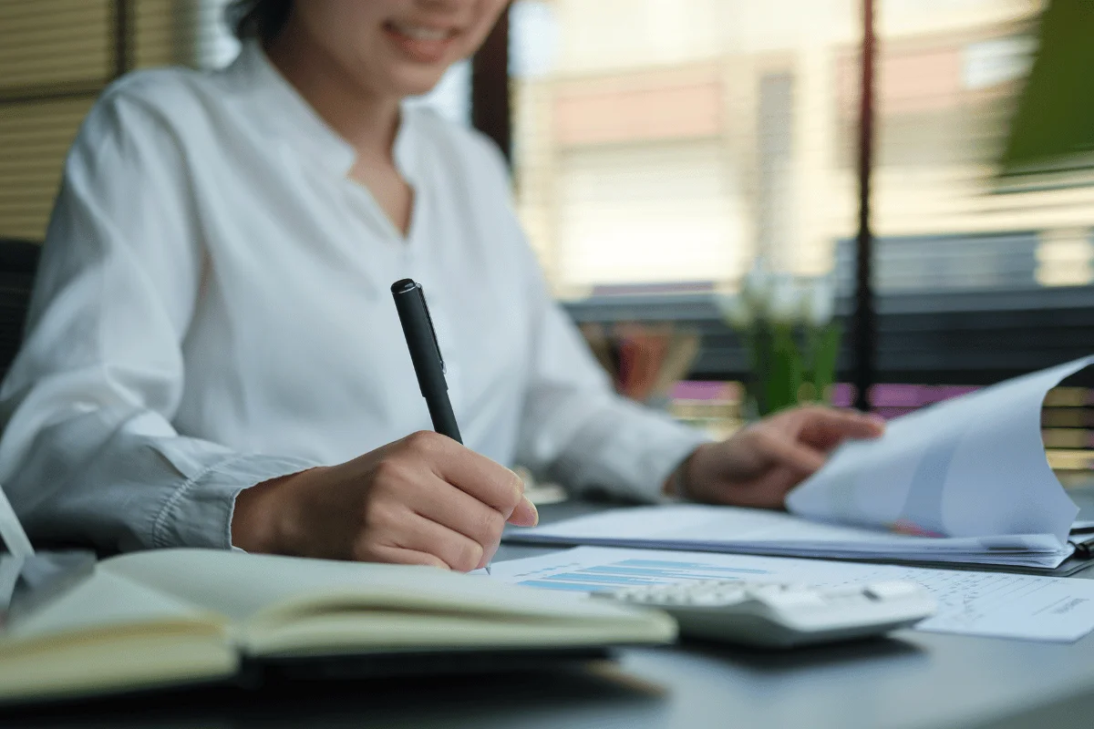 Businesswoman in a white blouse writing notes while reviewing financial documents at her office desk.