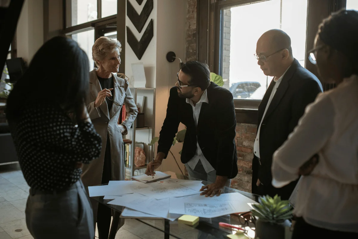 A group of professionals engaged in a collaborative discussion around a table covered with documents in a bright office setting.