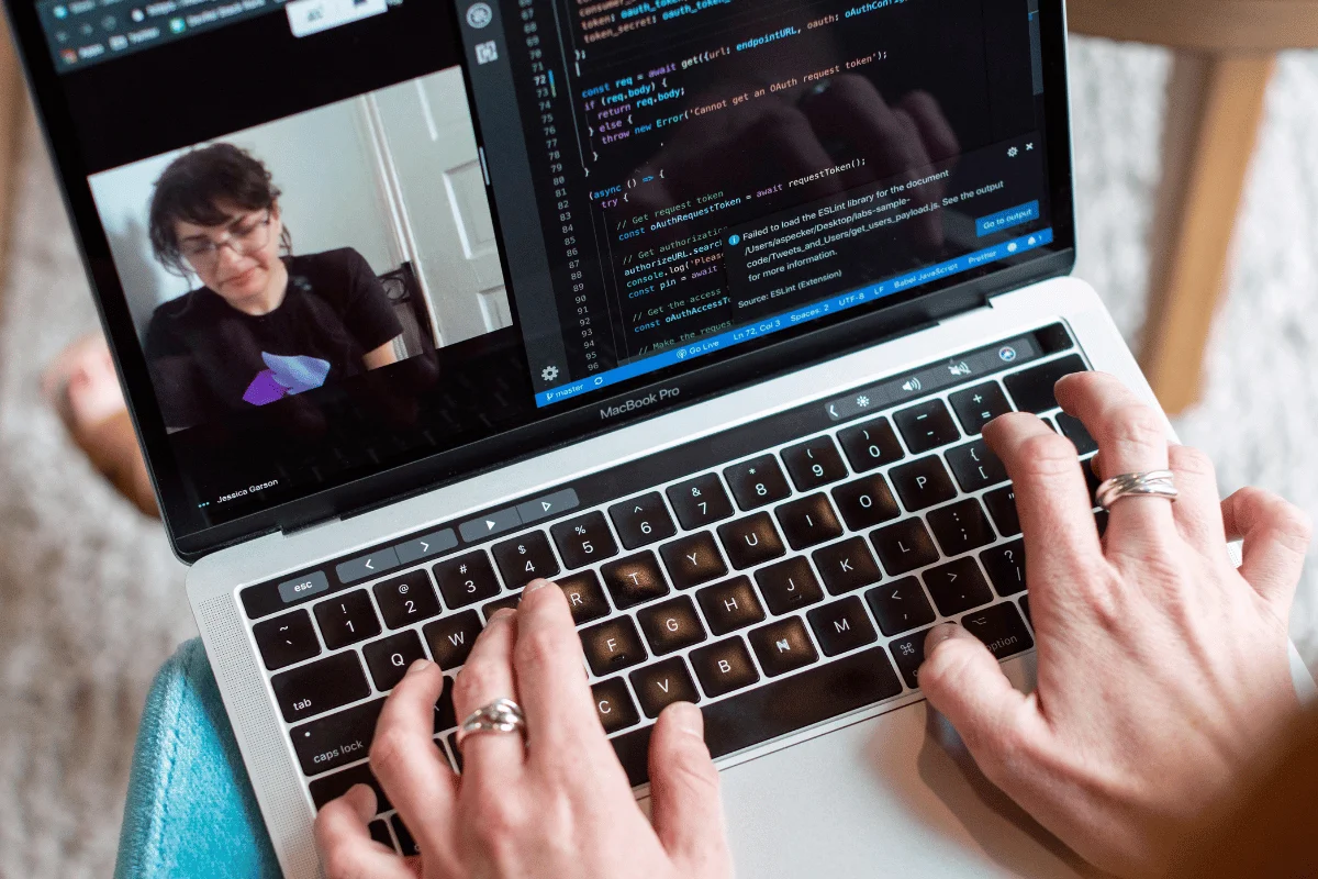 A person's hands typing on a MacBook Pro keyboard, with a coding interface displayed on the screen and a video call in progress.