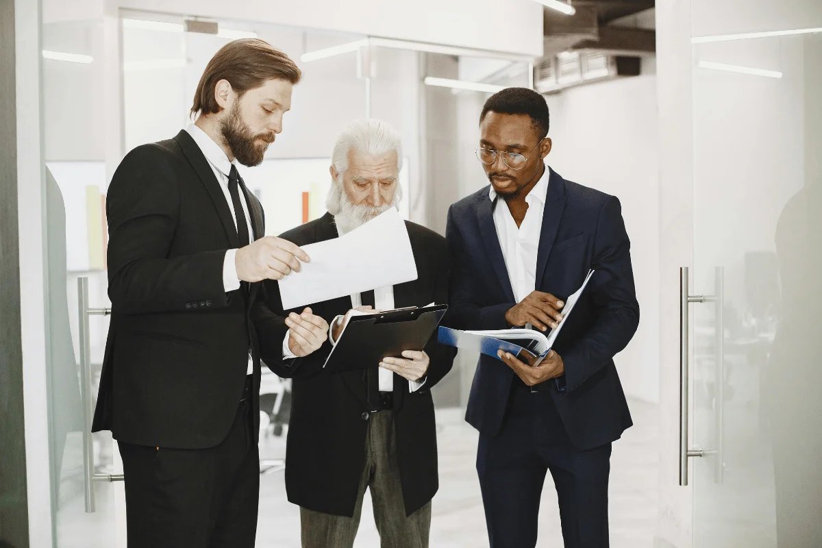 Three business professionals in formal attire engage in discussion while reviewing documents in a modern office setting.