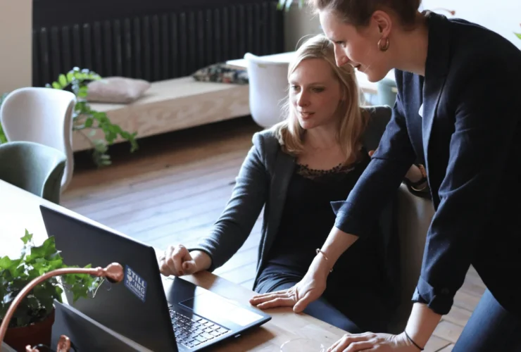 Two women engaged in a collaborative discussion at a desk, focusing on a laptop, with plants and a cozy workspace in the background.