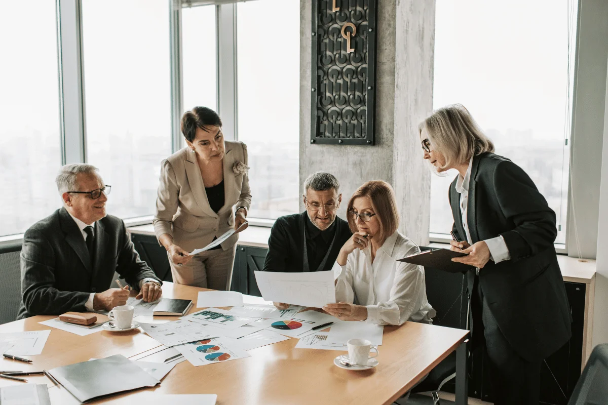 A group of five people in business attire gather around a table in a conference room, discussing documents and charts.