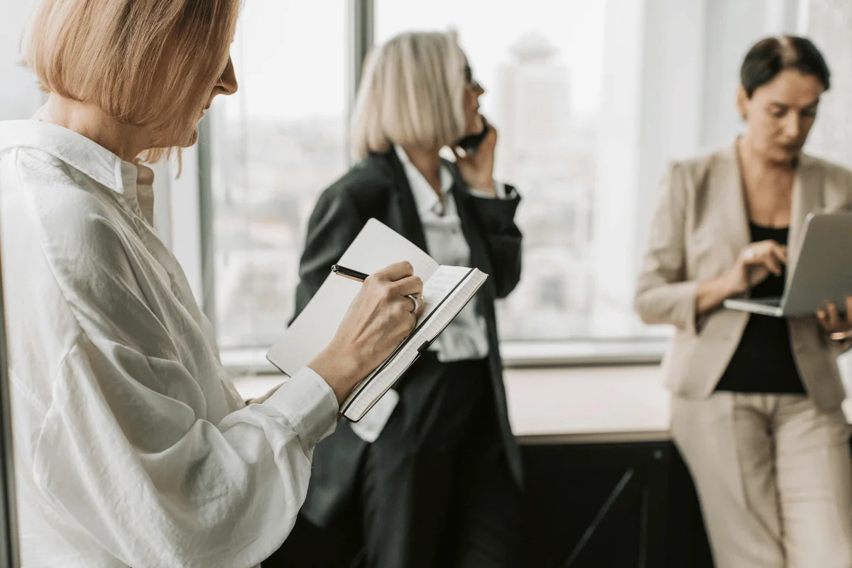 Three professional women in an office. One takes notes, another uses a phone, and the third works on a laptop. The mood is focused and productive.