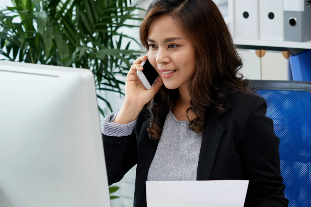Smiling woman in a black blazer on the phone, holding a paper, sits at a desk with a computer. Green plants and files in the background convey a professional office setting.