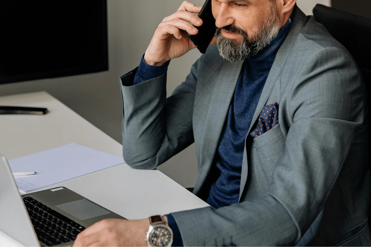 A man wearing a gray suit and blue shirt, talking on the phone while using a laptop.