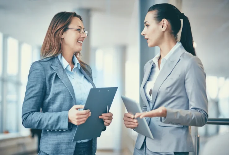 Two businesswomen in professional attire engage in a discussion, holding clipboards and a tablet in a modern office setting.