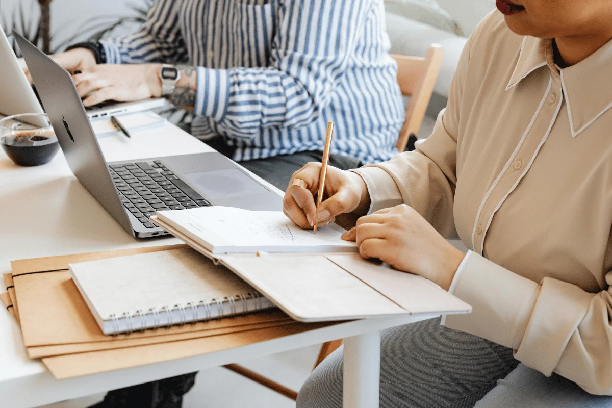 Two professionals collaborating at a desk, one writing in a notebook while the other works on a laptop.